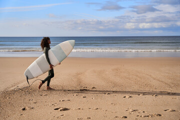 Cinematic shot of the curly woman surfer walking with surfboard on sandy coastline with footprints