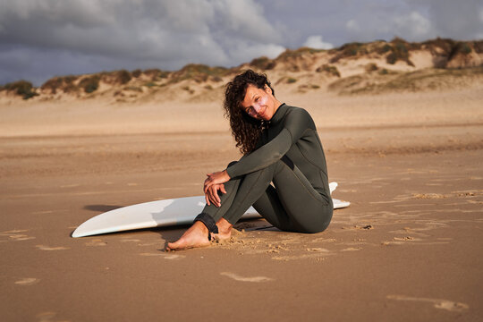 Positive female surfer looking at the camera while sitting with surf board on the sandy beach