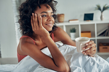 Dreamful African woman enjoying morning coffee while spending lazy time in bed