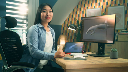 Female asian 3D designer sitting at the table near the modern computer and digital tablet, working on 3D project remotely from home and looking at camera.