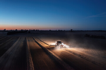 Panoramic aerial landscape view of working combine harvester at night with lights illuminating the...