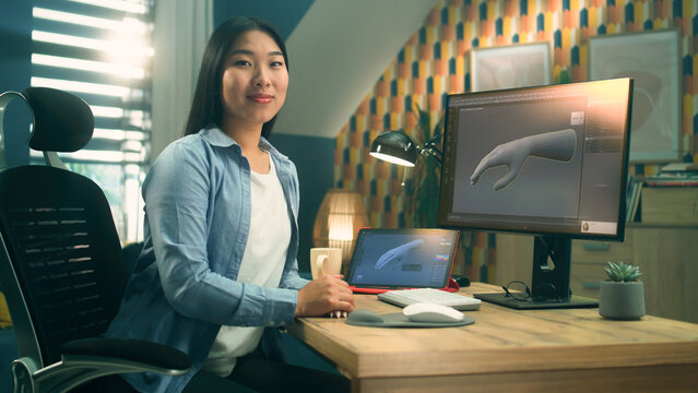 Female asian 3D designer sitting at the table near the modern computer and digital tablet, working on 3D project remotely from home and looking at camera.