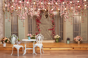 two white chairs and a table against the backdrop of decorations with pink flowers. photo zone with pink sakura flowers in a photo studio. decorative entrance to the house with furniture and a door.