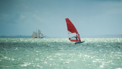 A man rides a hydrofoil windsurfer with a bright red sail on a beautiful turquoise green sea, blue sky, bright sunshine summer day, with an old tall ship on the horizon in the background.