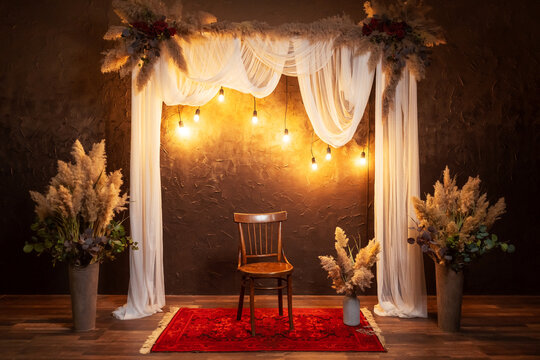 arch with white fabric against a brown wall with flowers. wedding classic arch with a chair for a photo shoot of weddings. wooden arch with dry flowers and incandescent lamps in a photo studio