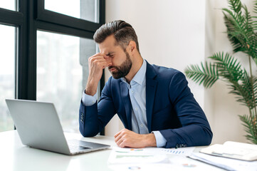 Naklejka premium Unhappy tired caucasian businessman, corporate manager, sits at the workplace, tired from online work, stressed, nervous, massages the bridge of his nose, closed his eyes, experiencing a headache