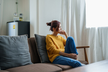 Obraz premium A portrait of a young Asian woman resting with a smiling face on the sofa by the window of the house