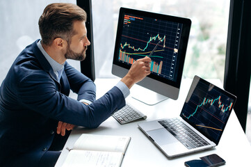 Focused smart successful caucasian male stock investor, broker, sits at his work desk, uses computer and laptop, concentrated analyzes risks and prospects, rise or fall of cryptocurrency coin