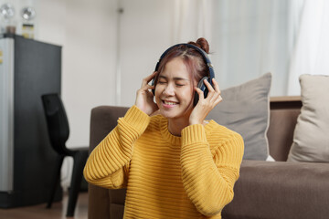 A portrait of a young Asian woman with a smiling face wearing a pair of headphones and using computer and listening to music while sitting on the sofa