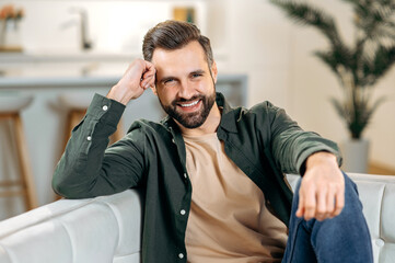 Male portrait. Headshot of a handsome attractive positive modern bearded caucasian man, in casual stylish clothes, sits on a sofa in the living room, looks at the camera, smiles friendly - obrazy, fototapety, plakaty