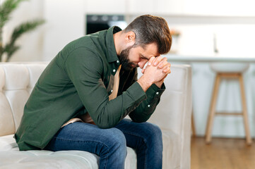 Upset frustrated caucasian man, sitting on the sofa in the living room with his head down and his eyes closed, experiencing personal problems, stress, loneliness, needs psychological support and rest