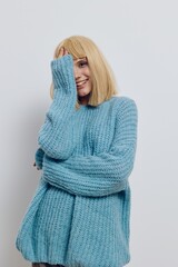 vertical photo of a beautiful, pleasant blonde woman in a long blue sweater with her hand covering her face, smiling pleasantly and looking at the camera