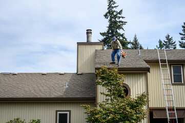 Senior man with a leaf blower on the roof of a residential home blowing debris out of gutters and off roof, fall maintenance
