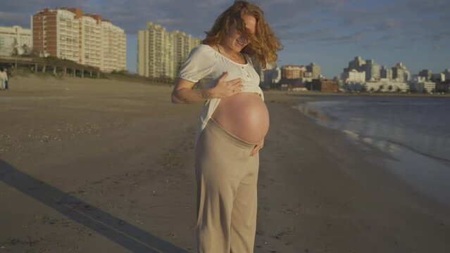 Pregnant caucasian redhead woman showing big belly while walking on beach at sunset, Punta del Este Uruguay. Handheld