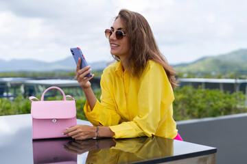 Stylish fit fashion women in bright yellow shirt trendy sunglasses posing at rooftop terrace tropical view outdoor holding mobile phone