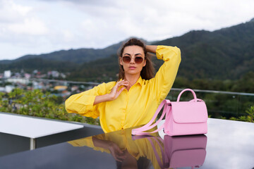 Fototapeta na wymiar Stylish fit fashion women in bright yellow shirt trendy sunglasses posing at rooftop terrace tropical view outdoor with pink bag