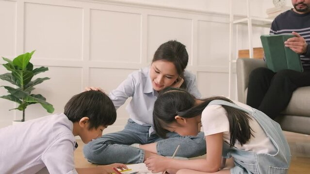 Asian Thai siblings and mum are sitting on living room floor, drawing with colored pencils together, dad leisurely relaxing on a sofa, lovely happy weekend activity, and domestic wellbeing lifestyle.