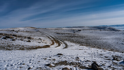 A dirt road runs along a snow-covered high-altitude plateau. Tire tracks bend and go to infinity. There are stones scattered around. A mountain range in the distance. Blue sky with radial clouds. 