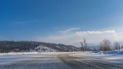 Obraz premium The highway curves through a snowy valley. Snowdrifts on the roadsides. Bare trees and a wooded hill against a blue sky. Altai. Copy space