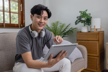 Fototapeta premium Handsome Asian man using digital tablet while headphones sitting on sofa at home