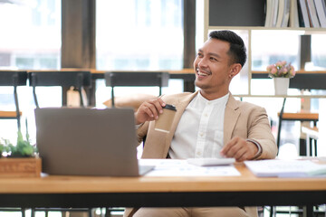 Young Asian businessman in suit and holding hot coffee cup and looking on laptop computer in office. Man in suit using laptop in well-lit workplace.