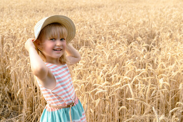 Child blonde girl in straw hat and nice dress smiling at the camera in wheat field
