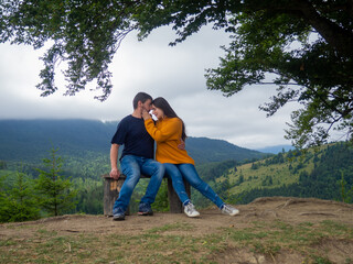 Young man and woman admiring breathtaking view while sitting on bench in the mountains. Lovely couple in casual clothes sit under large tree with forest background.