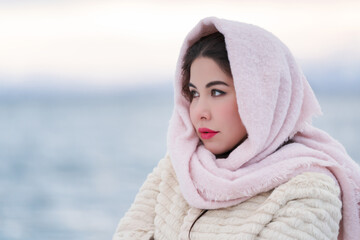 Obraz premium Portrait melancholy young woman on frosty winter evening outdoors. Elegant female with red plump lips in white fur coat and pink scarf on head looking away. Copy space. Depth of field, selective focus