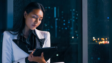 An Asian businesswoman using a tablet to work while standing by the window of an office late at...