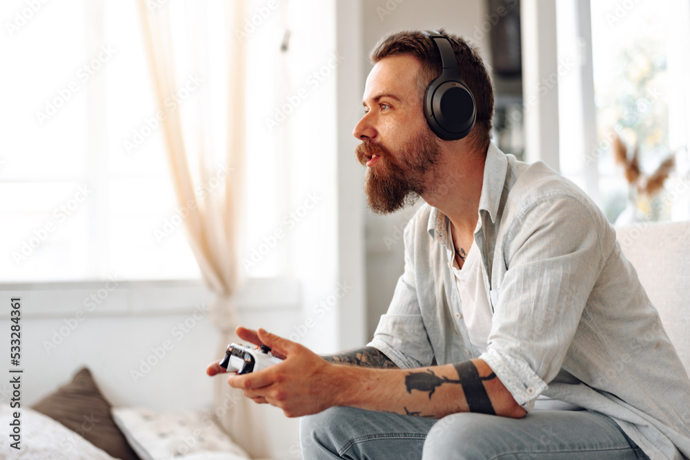 Wall mural Young bearded man playing video games while sitting on the couch at home