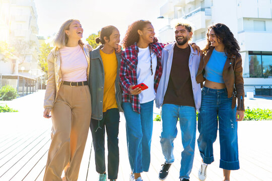 Group of young multiracial people walk with smartphones . happy group of multiethnic friends hugs laugh . College teenagers outdoors under the sun - friendship concept 