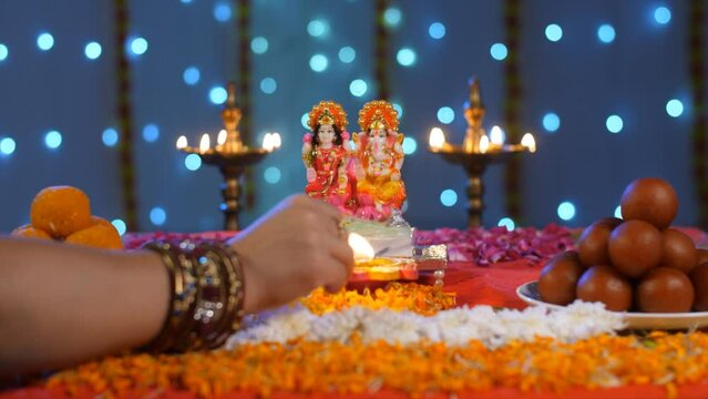 A female lighting a colorful Diya on the occasion of Diwali / Ganesh Chaturthi. Indian traditional sweets / Laddus  Gulaab Jamuns  brass Diyas  and idols of Lord Ganesha and Lakshmi Devi - a Hindu ...