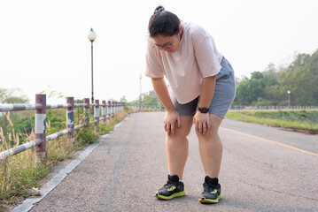 Wide shot portrait of overweight Asian woman wearing eyeglasses, standing outdoor, bending down, resting after running, being tired, suffering from knee and joint injury. Sport and obesity concept.