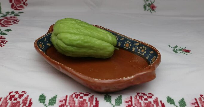 Adult female hands placing two chayotes in a clay tray on table, cross-stitch tablecloth, labu siam, jipang, organically grown vegetables. Concept of bio, biological and natural cultivation at home