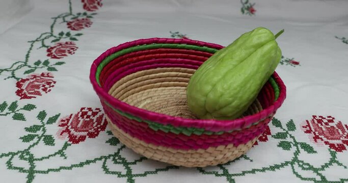 Female hands placing two chayotes in a Mexican wicker basket on a tablecloth with cross-stitch flowers, labu siam, jipang organically grown vegetables. Concept of bio, biological, cultivation at home