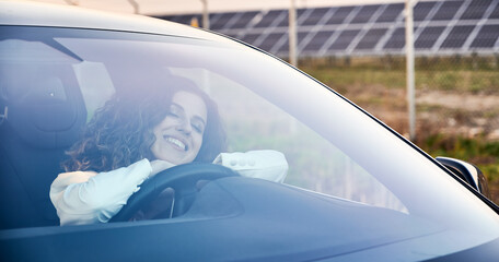 Close-up view of happy woman placing chin on folded arms at wheel of car. Girl enjoying favorite vehicle, closed eyes against solar panels. Concept modern use of electricity in automotive industry.