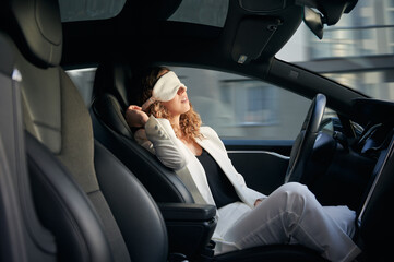 Woman in sleeping mask taking nap in electric car with vehicle autopilot system. Stylish businesswoman sleeping in electro car while using driver-assistance system.