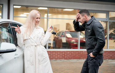 Happy blonde woman showing keys to newly bought white car to her surprised boyfriend outdoors on background of automobil dealership. Anxious man covering his forehead with hand, looking to new buy.