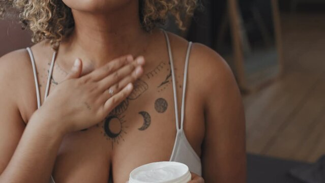 Slow motion close-up shot of young Black woman sitting in loft bedroom in morning applying and dabbing moisturizing cream over her face and neck