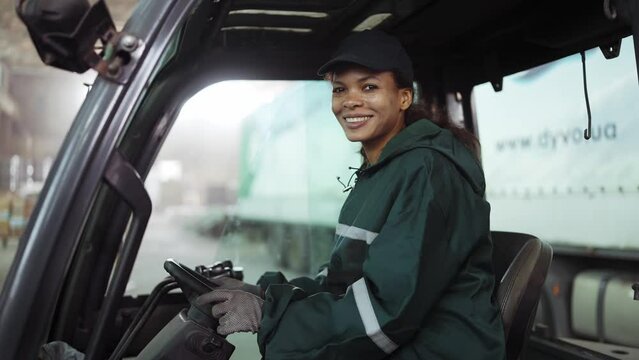 Potrait of an African American woman in a special green uniform sitting behind the wheel of a truck in a waste processing plant. Processing of raw materials, recycling. Pollution control