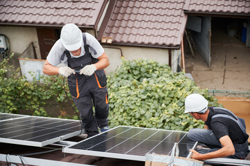 Men workers mounting photovoltaic solar moduls on roof of house. Electrician in helmets wiring...