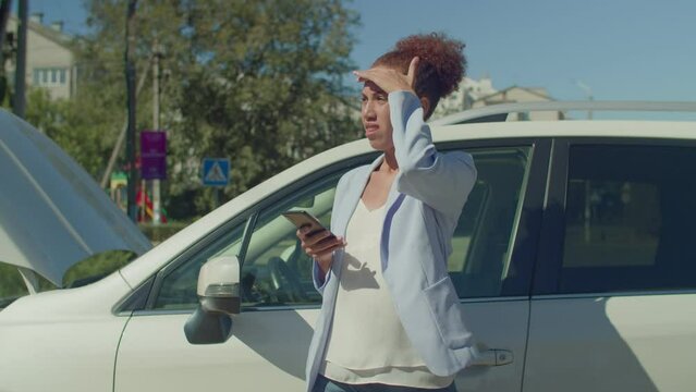 Worried nervous charming black woman driver standing near broken vehicle, browsing online using smartphone and looking for roadside assistance service while travelling by car on business trip.