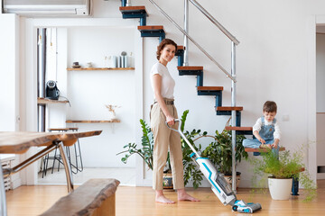 woman with cordless vacuum cleaner cleans floor, preschool child on appartment