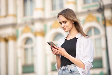 A young and attractive Caucasian girl in casual clothes smiles and uses a mobile phone in the city on a sunny day.