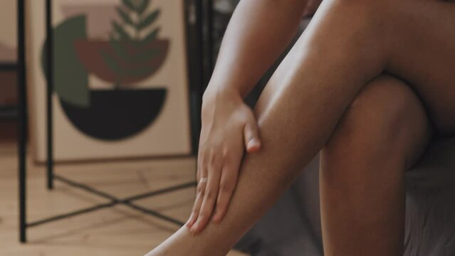 Attractive young African American woman with tattoo on chest sitting on bed in morning applying and dabbing body cream over her legs