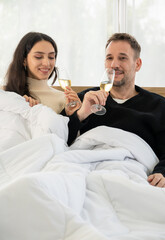 Couple celebrating holiday at home together, sitting on bed in sweaters and drinking champagne.