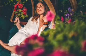 beautiful woman sits on a wooden swing with a bouquet of red roses in her hand.