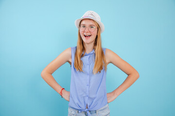 Portrait of teenage girl with open mouth wearing white summer hat, glasses, holding hands on waist on blue background.