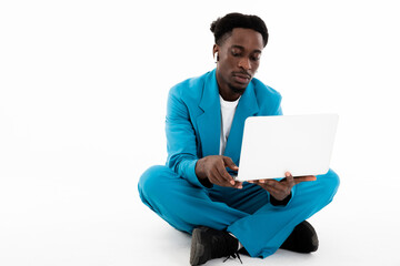 Serious african american man sitting on floor on white background in studio isolated looking at screen holding modern laptop listening to music by modern earphones.