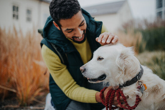 Happy young man stroking his dog outdoors in city park, during cold autumn day.